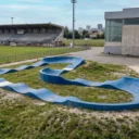 circuit de pumptrack Y1 vu de profil aux abords d'un terrain de rugby et d'un gymnase